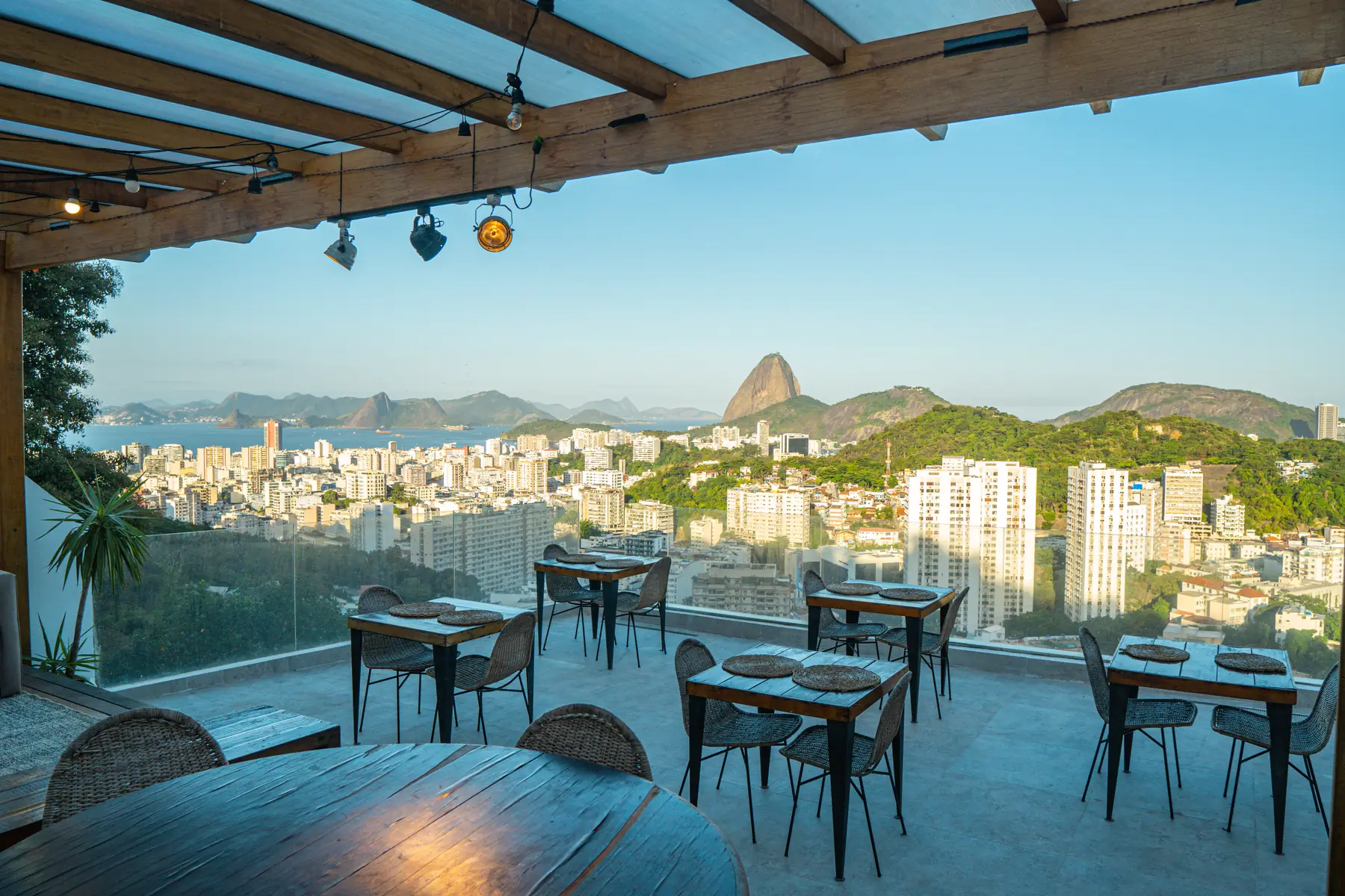 Rooftop ao entardecer, com luzes na pérgola, mesas postas e o Pão de Açúcar ao fundo