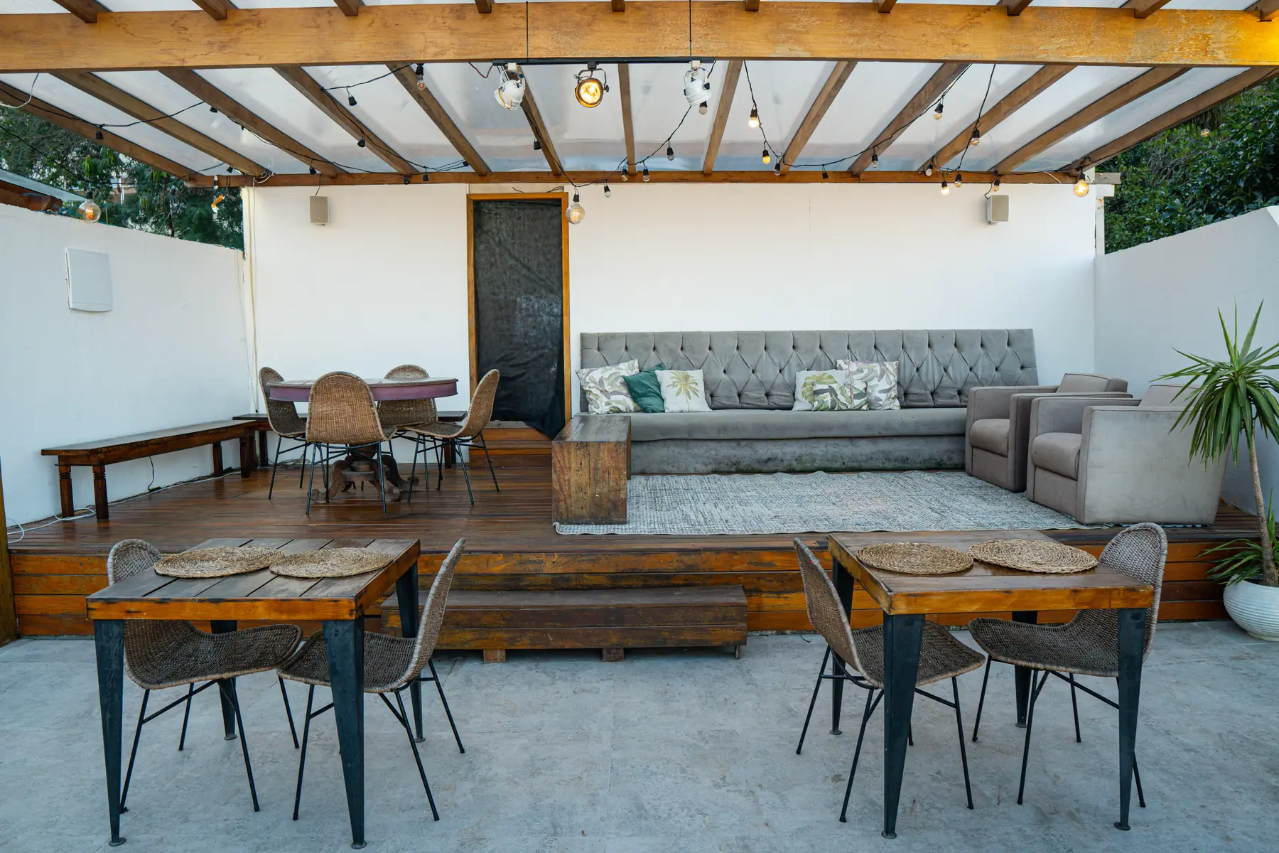 Área externa com mesas e cadeiras de madeira, sofá cinza com almofadas decorativas e poltronas sob cobertura de madeira com iluminação suspensa.